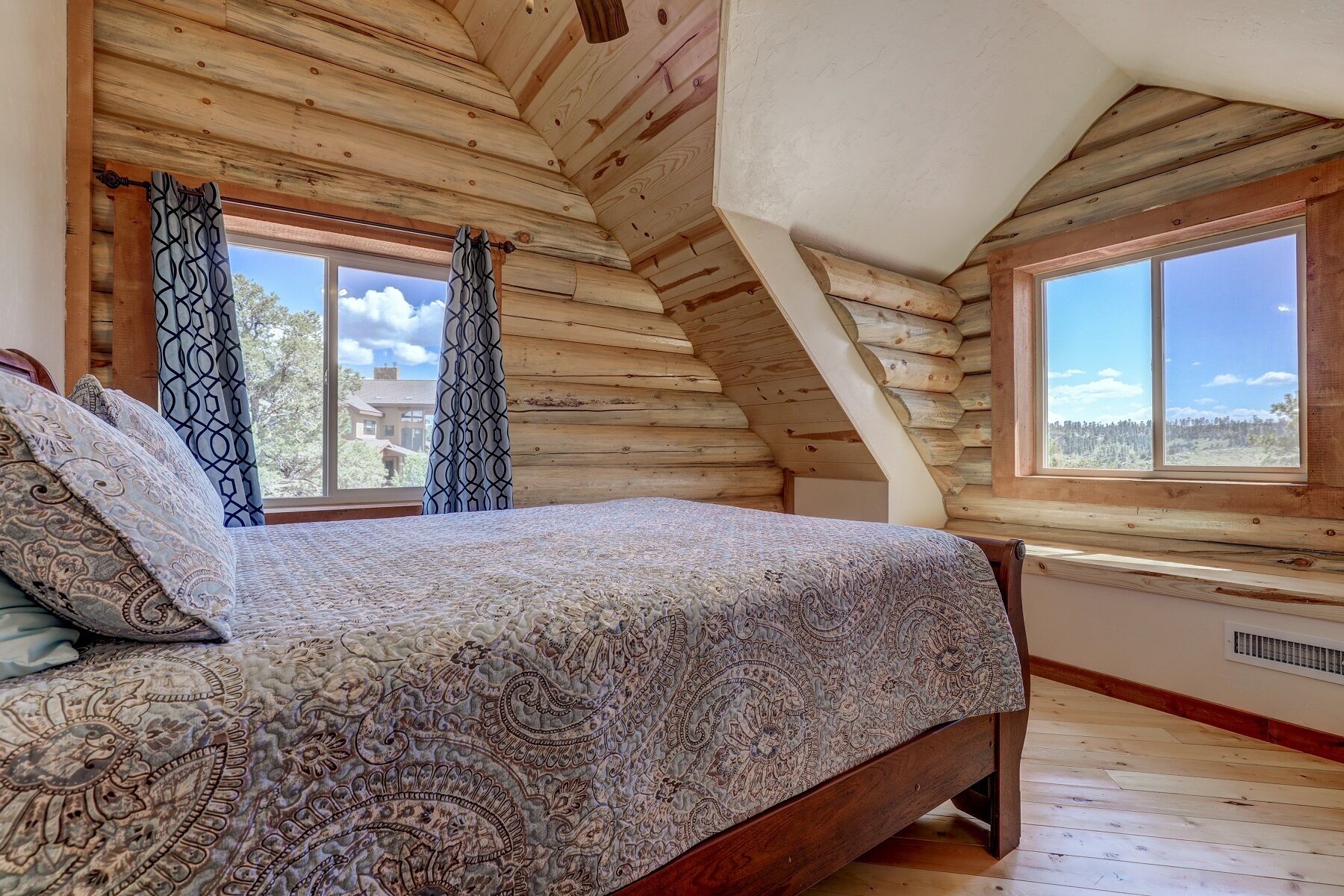 A bedroom with a wooden sloped ceiling, a large bed with patterned bedding, two windows with curtains, and a view of a forest.