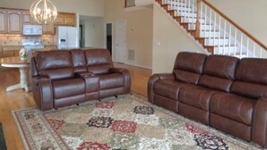 A spacious living area with two brown leather sofas, a patterned rug, a chandelier, and a staircase leading to an upper floor.