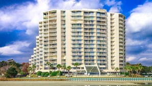 A high-rise hotel with multiple balconies, a reflective pool in the foreground, and palm trees in the background.