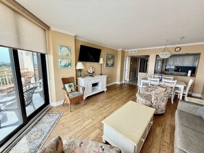 A spacious living area with a large window, a dining table, a sofa, and a TV mounted on the wall. - South Tower JJ4- Oceanfront-What a View! Remodeled Kitchen & Baths! (Myrtle Beach)