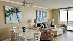 A living room with a dining table, white chairs, a grey sofa, and a patterned armchair. There are two paintings on the walls, a ceiling fan, and a large window with a view.