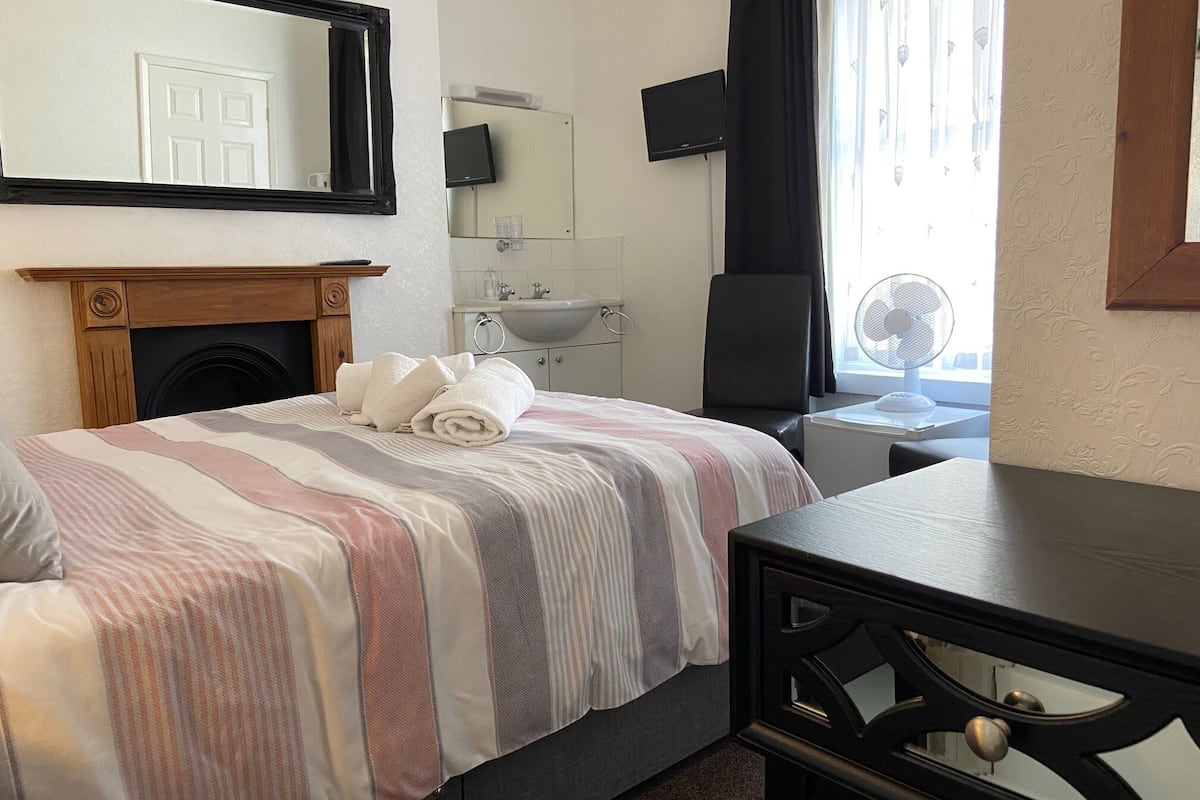 A hotel room with a bed, a wooden dresser, a desk, a television, a mirror, a window with curtains, and a bathroom with a sink and mirror.