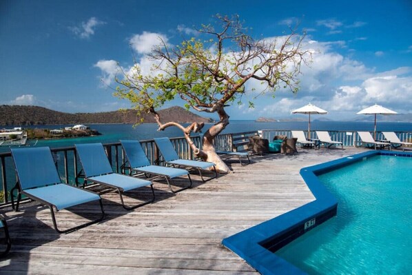 A wooden deck with lounge chairs and a pool, overlooking a body of water and hills.