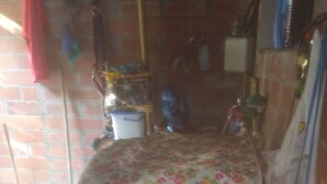 A room with a checkered floor, a bed covered with a floral blanket, a brick wall, and a window with a curtain. - Casa Wilber (Cali)