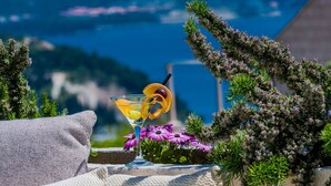 Ein gemütlicher Sitzbereich im Freien mit Blick auf Meer und Berge, mit einem Getränk, Kissen und einem Hut.