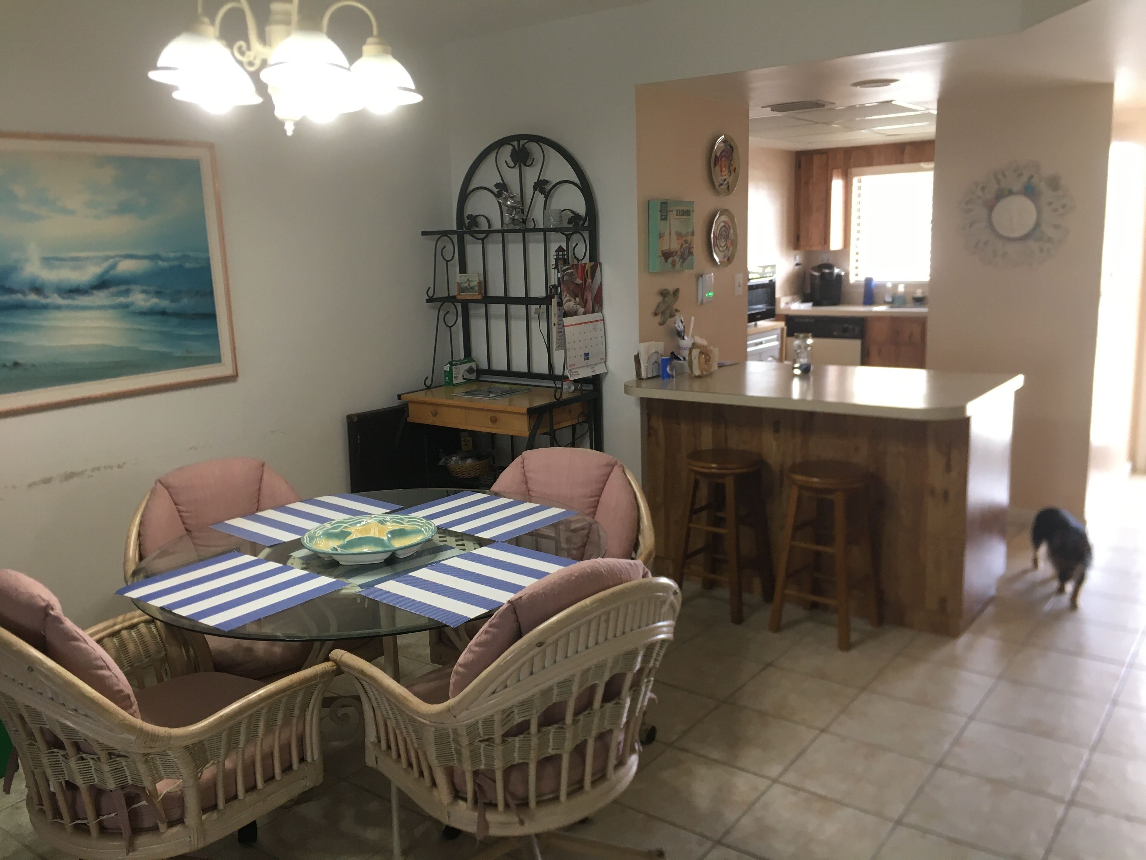 A dining area with a round table, striped tablecloth, wicker chairs, and a kitchen in the background.