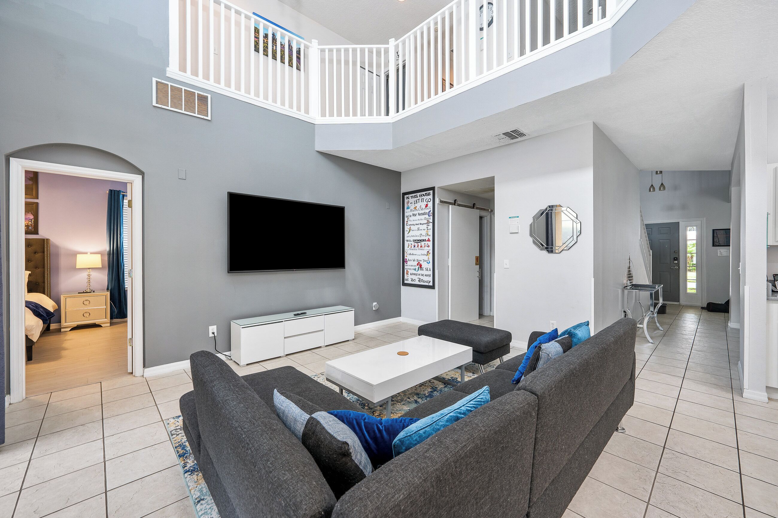 A modern living room with a flat-screen TV, grey sofas, a white coffee table, and a staircase leading to an upper level.