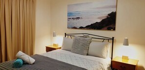 Una cama bien hecha con un edredón estampado, dos lámparas de noche, una mesita de noche con una lámpara, una toalla enrollada y un cuadro con una escena de playa en la pared.
