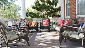 A covered patio with wicker furniture and cushions, a brick wall, and a view of trees. - Port City Guest House (Wilmington)