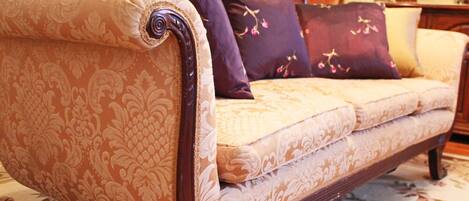 A vintage sofa with ornate patterns, a wooden armrest, and multiple cushions in a room with floral carpet and wooden furniture.