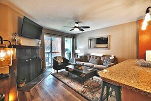 A modern living room with a fireplace, a television, a ceiling fan, and a kitchen area with a granite countertop. - Mountain Views! Hot Tub/Pool Walk to Lifts & Main1 (Park City)