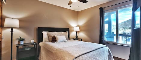 A hotel room with a bed, bedside lamps, a ceiling fan, a window with a view, and a small table with a plant.