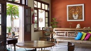 A room with a wooden table, chairs, a bench with colorful cushions, a large window with a view of greenery, and a lion painting on the wall. - Coqueiros Praia Hotel - Balneário de Iriri (Anchieta)
