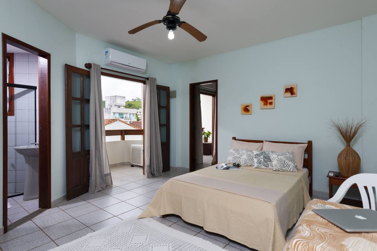 A bedroom with a bed, a ceiling fan, an air conditioner, a window with curtains, and a view of buildings outside.