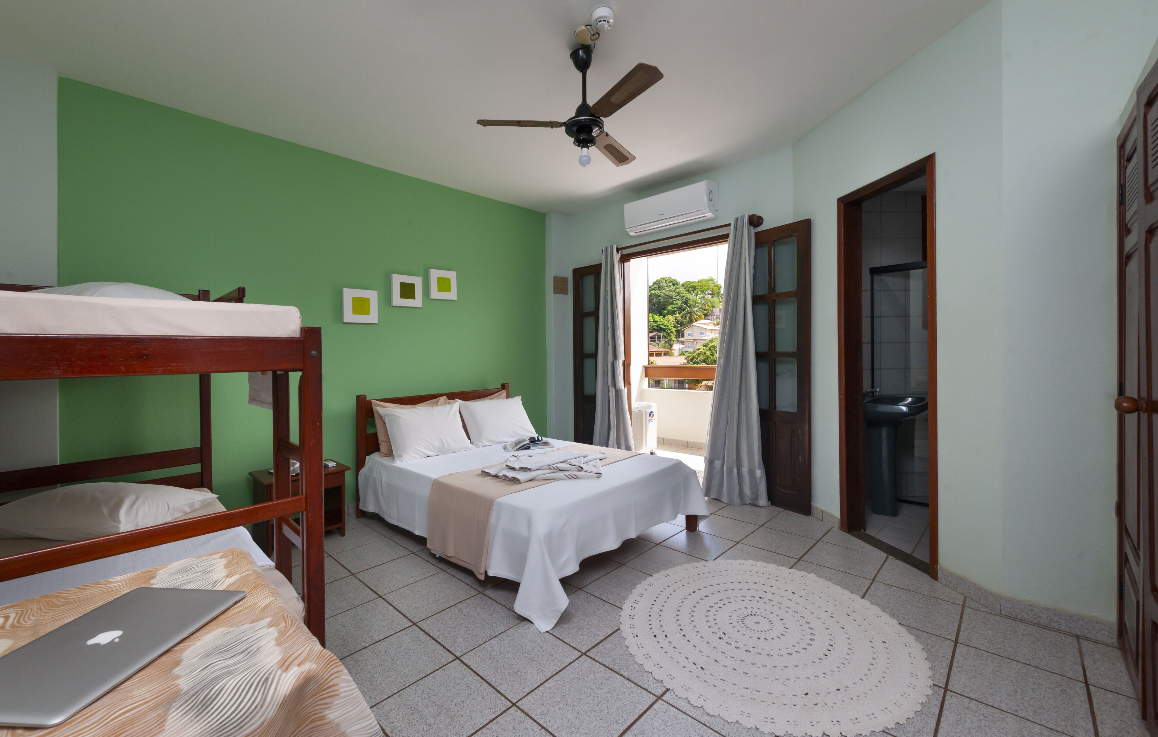 Quarto com beliches, uma cama de solteiro, um laptop sobre uma mesa, um ventilador de teto e vista para o exterior através de uma janela.
