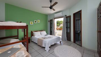 Quarto com beliches, uma cama de solteiro, um laptop sobre uma mesa, um ventilador de teto e vista para o exterior através de uma janela.