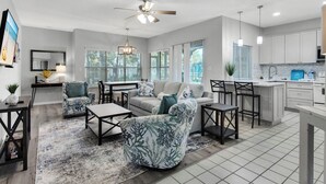 A spacious living room with a sofa, coffee table, and chairs, a dining area with a table and chairs, and a kitchen with white cabinets and a countertop. - Hidden Dunes Cottages 2-Bedrooms (Miramar Beach)