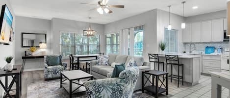 A spacious living room with a sofa, coffee table, and chairs, a dining area with a table and chairs, and a kitchen with white cabinets and a countertop.