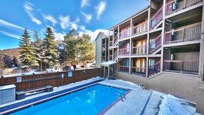 A swimming pool adjacent to a multi-story building with balconies, surrounded by trees and a wooden fence.