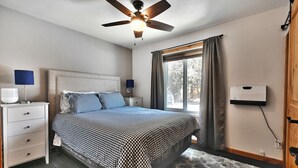 A bedroom with a bed, a ceiling fan, a nightstand with a lamp, a window with curtains, and a wall-mounted device.