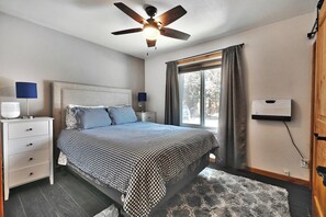 A bedroom with a bed, a ceiling fan, a nightstand with a lamp, a window with curtains, and a wall-mounted device. - First Chair Snowcrest Base Camp | Walk to Lifts (Park City)