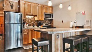 A modern kitchen with wooden cabinets, a stainless steel refrigerator, a built-in microwave, and a granite countertop. There are two bar stools and a small island with a sink.