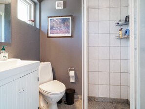 Un baño con inodoro, lavamanos y zona de ducha. Se aprecia un cuadro en la pared y artículos de aseo en la estantería.