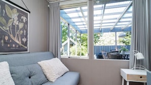Quarto com sofá cinzento, mesinha de cabeceira branca, impressão botânica emoldurada e janela com vista para vegetação e uma casa.