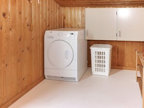 A laundry room with a front-loading washing machine, a white laundry basket, and wooden cabinets. - Cozy Holiday Home in Hadsund near Sea (Hadsund)