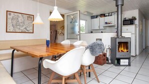 A cozy kitchen with a central wood-burning stove, white chairs, a wooden table, and hanging lights.