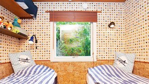 A room with two beds, a window with a view of greenery, patterned wallpaper, and a shelf with plush toys.