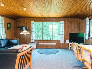 A room with a wooden ceiling, a television, a sofa, a dining table with chairs, and a window with a view of trees.