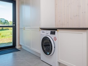Une buanderie équipée d’un lave-linge à chargement frontal, d’une porte donnant sur un espace verdoyant et de meubles blancs.