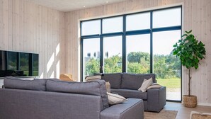 Een moderne woonkamer met grijze banken, een flatscreen televisie en een groot raam met uitzicht op groen.