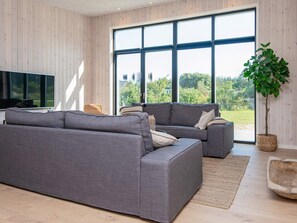 A modern living room with a grey sectional sofa, a flat-screen TV, and a large window offering a view of greenery. - 5 Star Holiday Home in Glesborg (Glesborg)