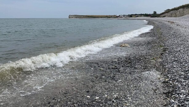 Een kiezelstrand met golven, een verre klif en een heldere lucht.
