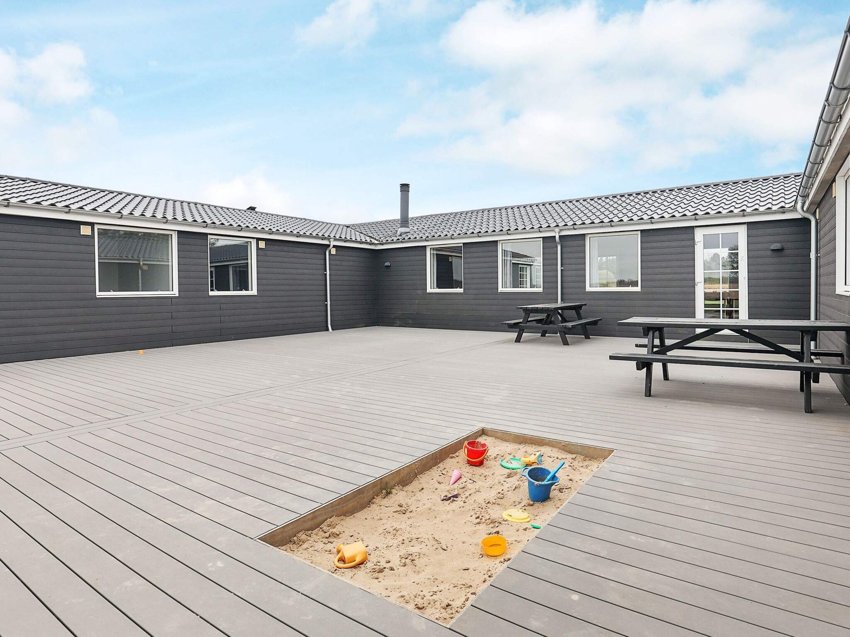 Een overdekt buitengebied met een houten terras, picknicktafels en een zandbak.
