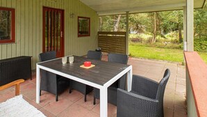 Alpendre coberto com mesa e cadeiras, uma porta vermelha e vista para o jardim.