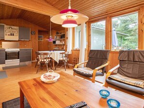 A cozy cabin interior with wooden walls, a dining area with a table and chairs, a sofa, a kitchen with a stove, and a large window offering a view of the outdoors.