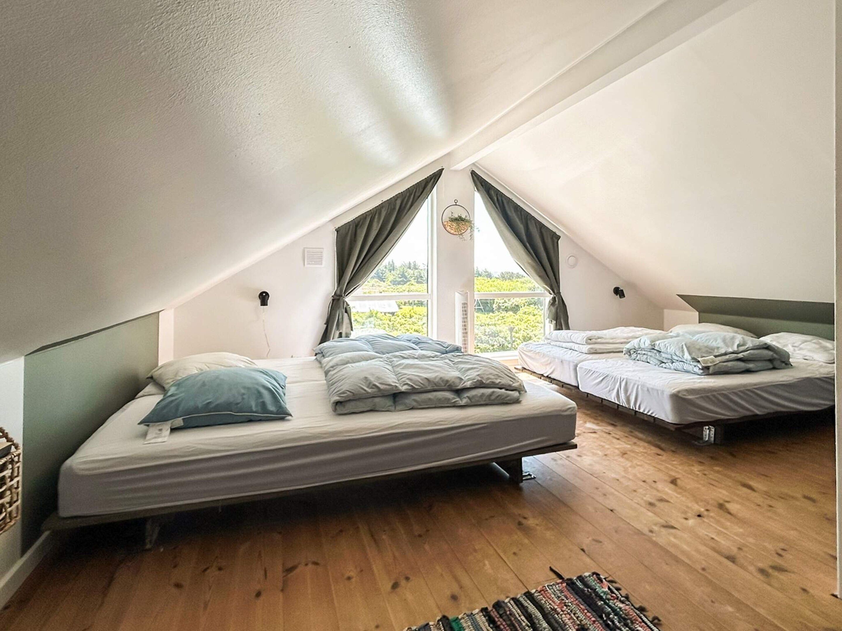 A small attic bedroom with two beds, wooden flooring, and a window with curtains.