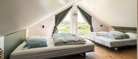 A small attic bedroom with two beds, wooden flooring, and a window with curtains.