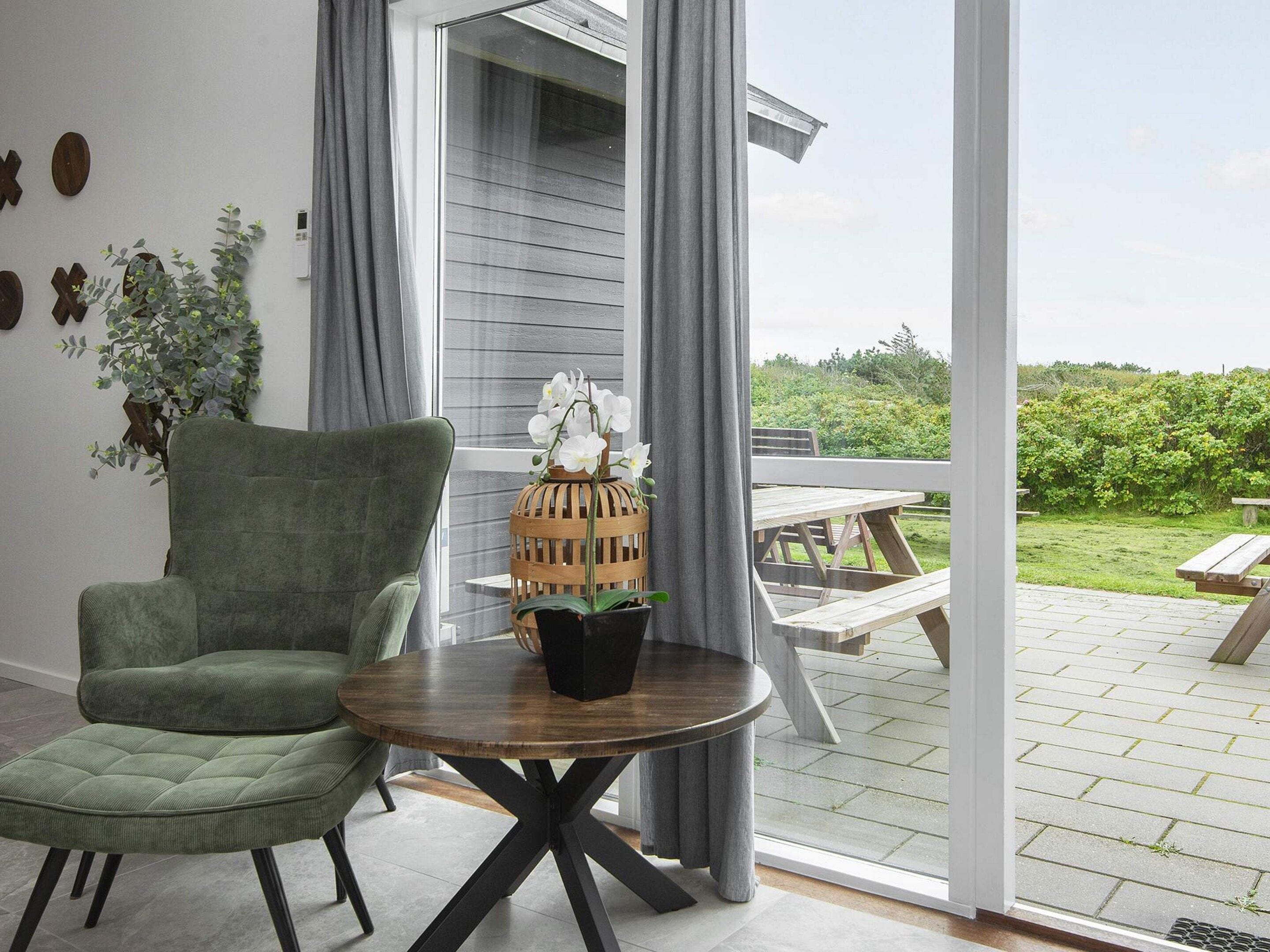 Et værelse med en grøn polstret stol, et træbord med en potteplante, og udsigt til en udendørs terrasse med et træbord og bænke.