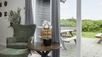 Et værelse med en grøn polstret stol, et træbord med en potteplante, og udsigt til en udendørs terrasse med et træbord og bænke.