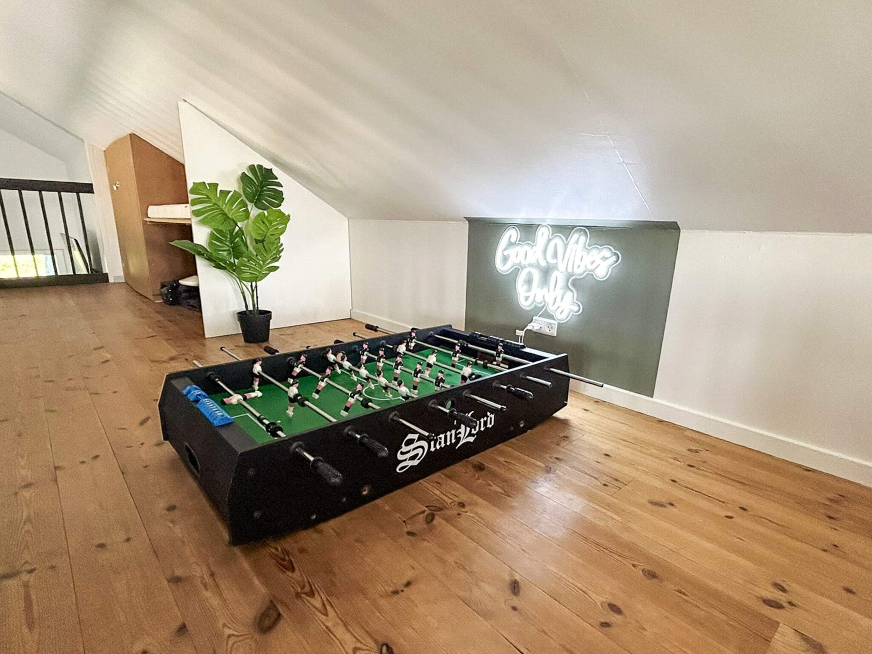 Una mesa de fútbol de mesa en una habitación con piso de madera, una planta en maceta y un letrero de neón en la pared.