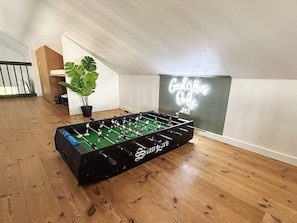Una mesa de fútbol de mesa en una habitación con piso de madera, una planta en maceta y un letrero de neón en la pared.
