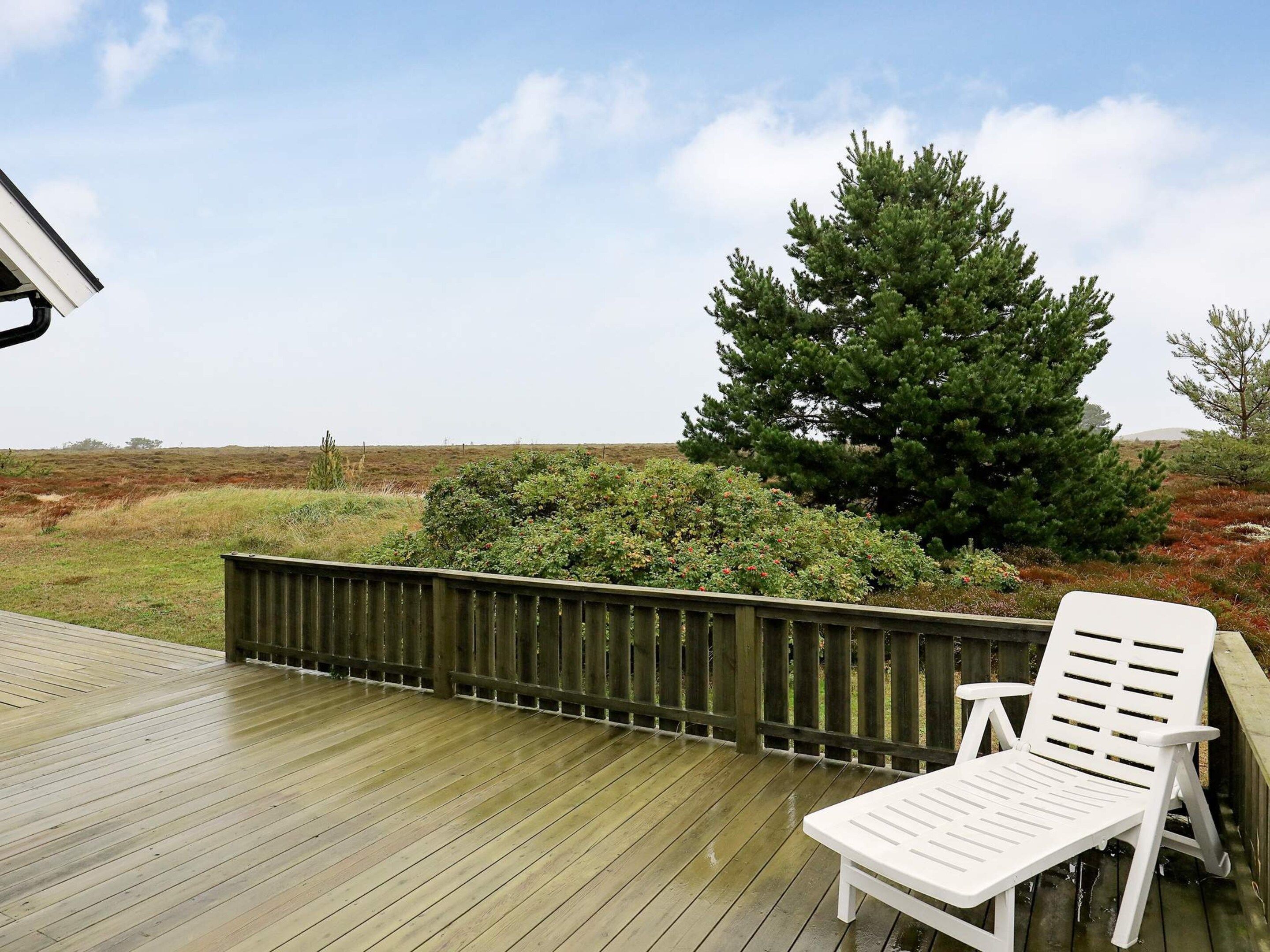 A wooden deck with a white lounge chair, a wooden fence, and a large coniferous tree in the background.
