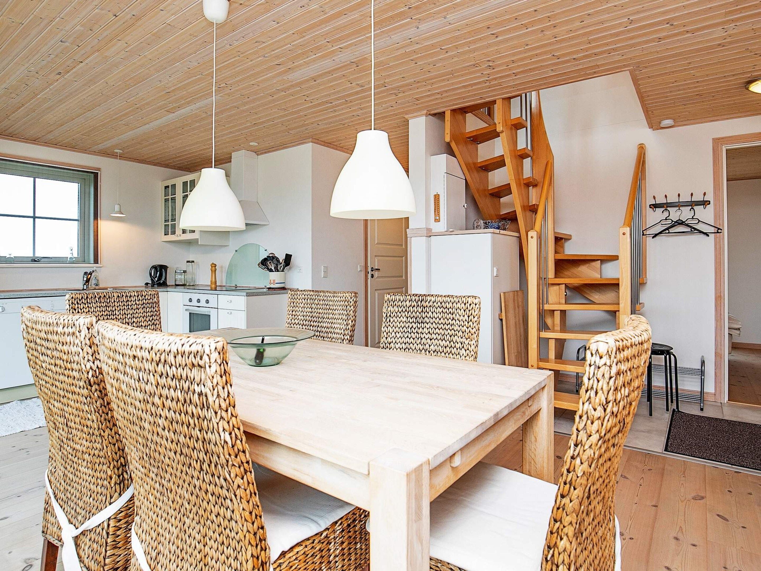 A modern kitchen with white cabinets, a wooden staircase, and a dining area with wicker chairs and a wooden table.