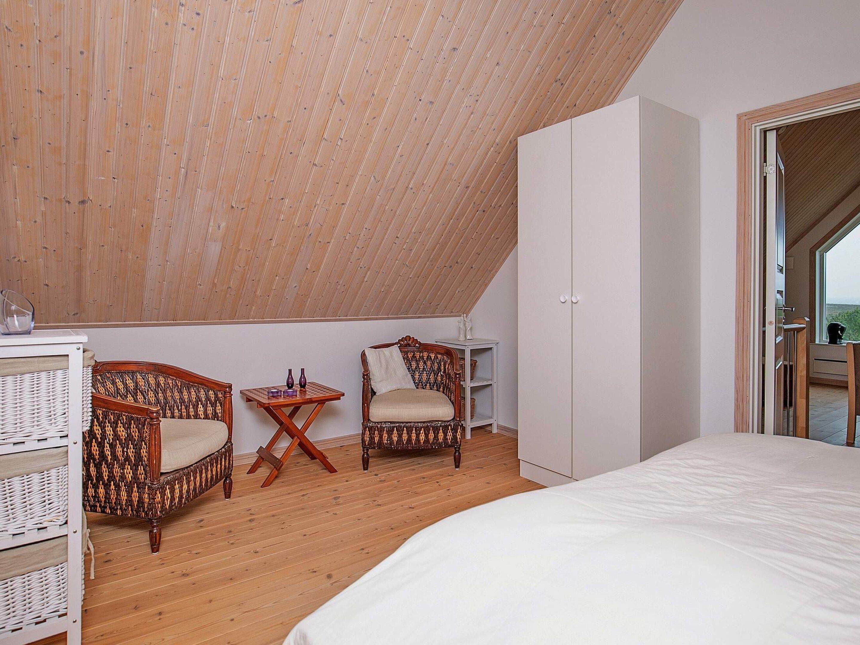 Ein Schlafzimmer mit geneigter Decke, zwei Korbstühle, ein kleiner Tisch mit Dekoration, ein Kleiderschrank und ein Bett.
