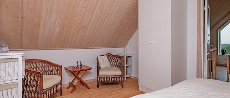 A bedroom with a sloped ceiling, two wicker chairs, a small table with decorative items, a wardrobe, and a bed.