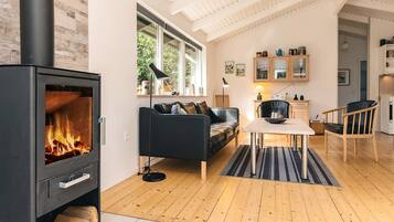 A modern living room with a wood-burning stove, a sofa, a dining table, and a kitchen area with cabinets and a refrigerator.
